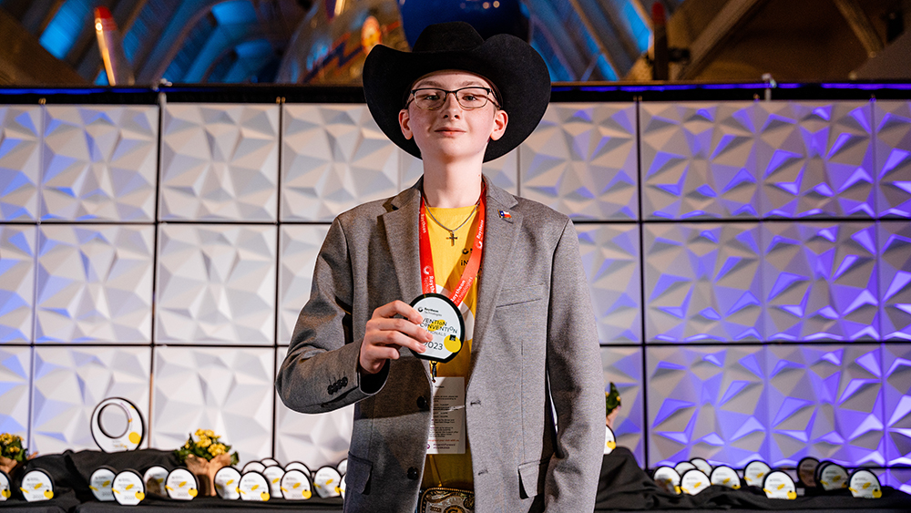 Young male student wears a black cowboy hat and sports coat while looking at the camera and holding up an award medal. 