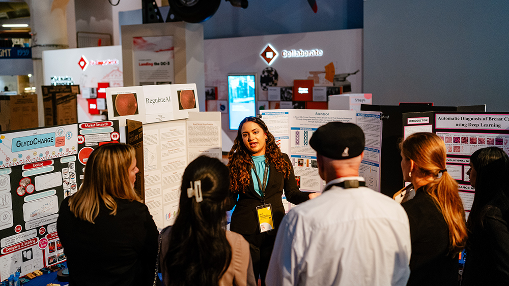 Female student wearing a teal shirt and black blazer stands in front of a project board while speaking to adults standing in front of her with their backs to the camera. 