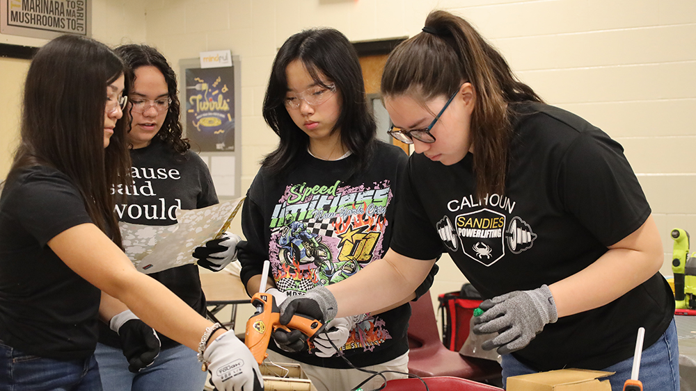 Four high school students wearing protective eyewear and gloves stand around a table working on an engineering design project with a hot glue gun. 