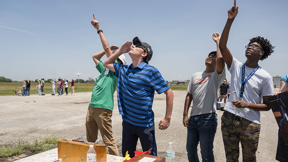 Students stand outside looking up and pointing to the sky as they watch their model rockets soar into the sky. 