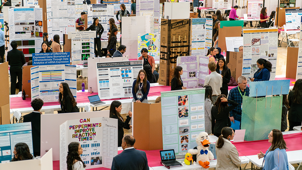 Overhead camera shot of students and adult judges in a gym with tables full of science fair posters.