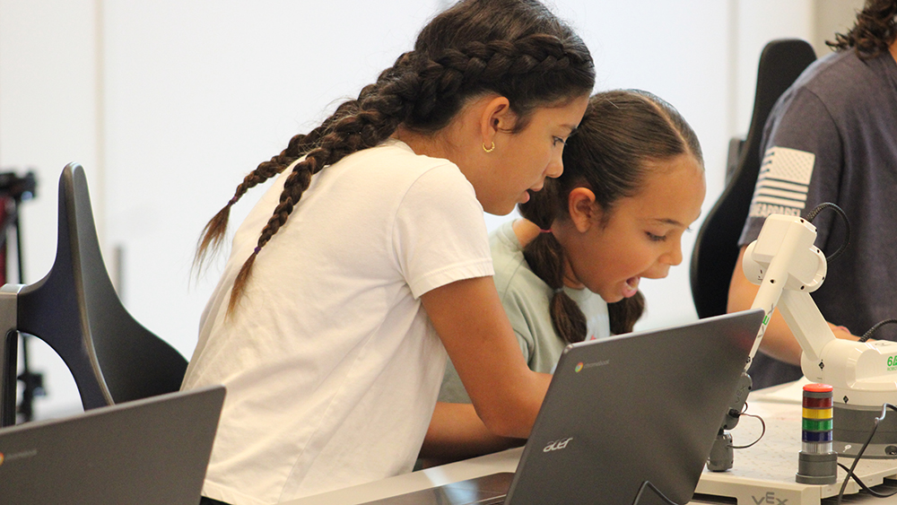 Two elementary-age girls work at a classroom desk using a laptop computer to program a small robot arm. 