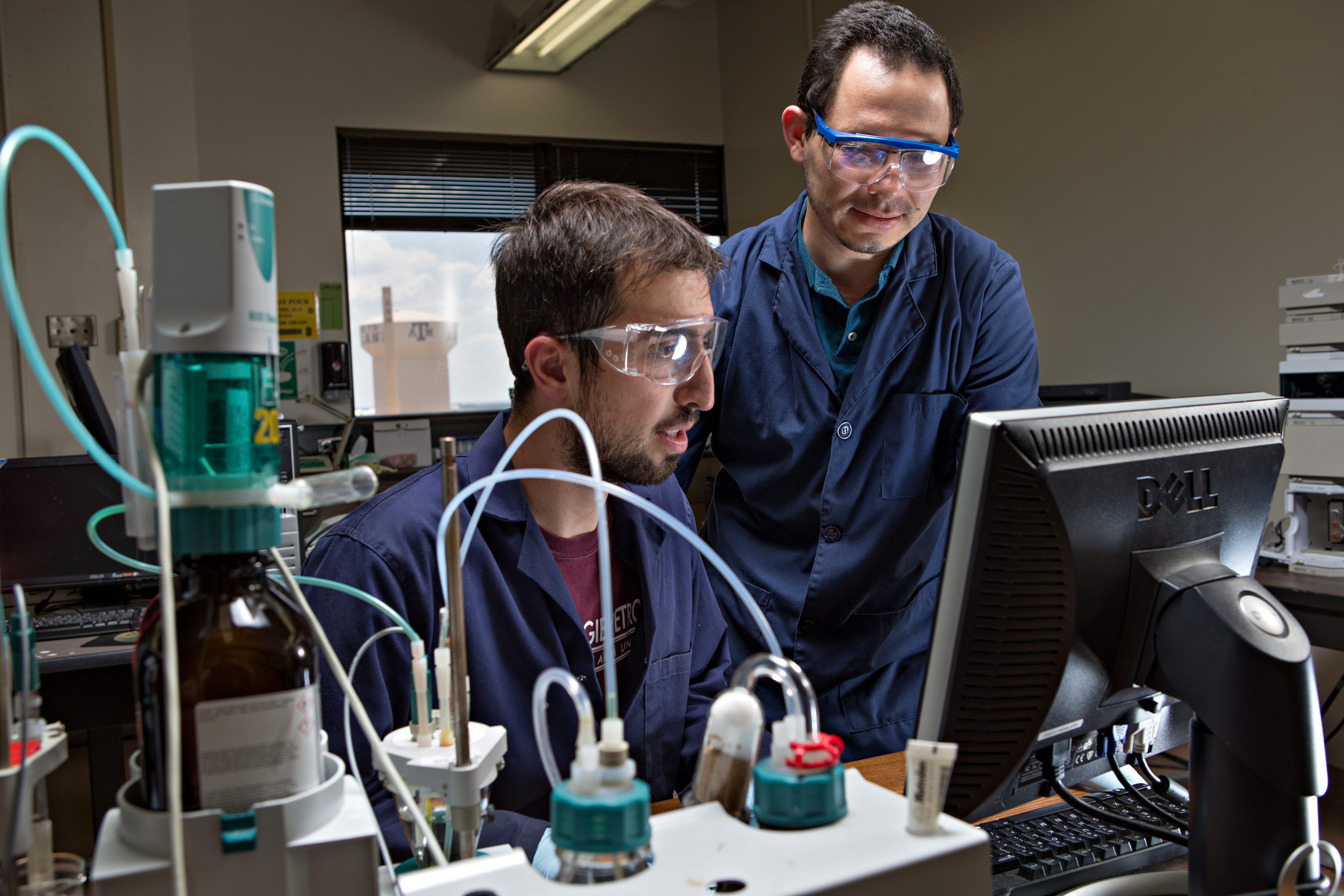 Two male students in navy lab coats stand in front of a computer in a lab. There is an open window in the background that shows a water tower. 