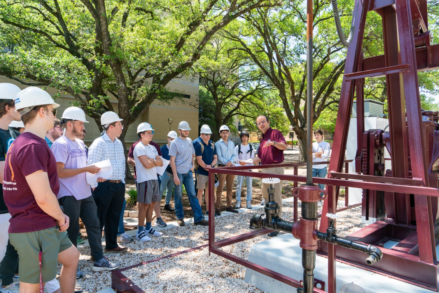 A group of students in hard hats stand in front of an oil pump during a class. Trees and various buildings are in the background. 