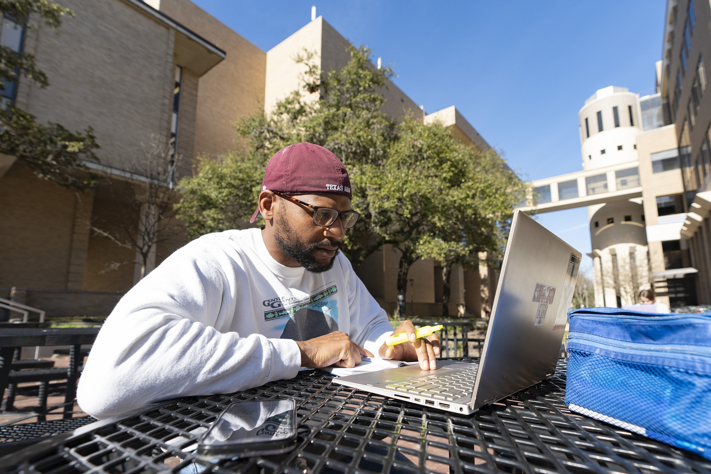 A male student sits at a table outdoors, wearing a backwards A&amp;M hat. He is holding a highlighter and working on a laptop. 