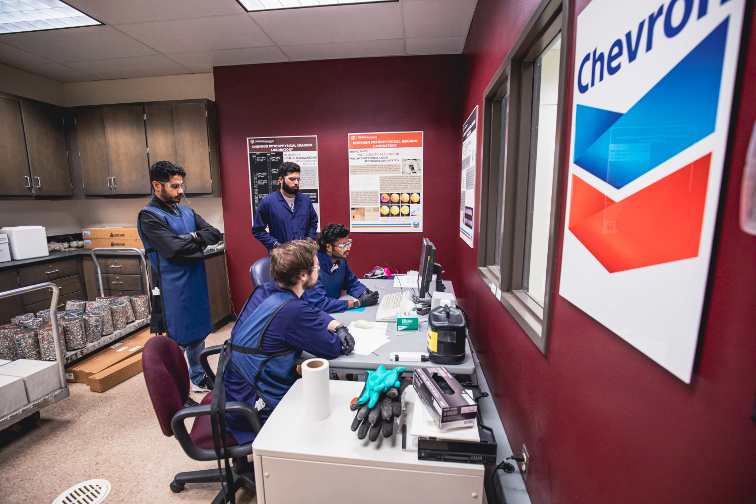 Four students are in a lab; two are standing and two are sitting. They are seated in front of a computer that they are each looking at. 