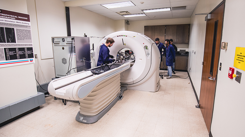 Several students in lab coats and safety gear working around a computerized tomography scanner centered in a large room