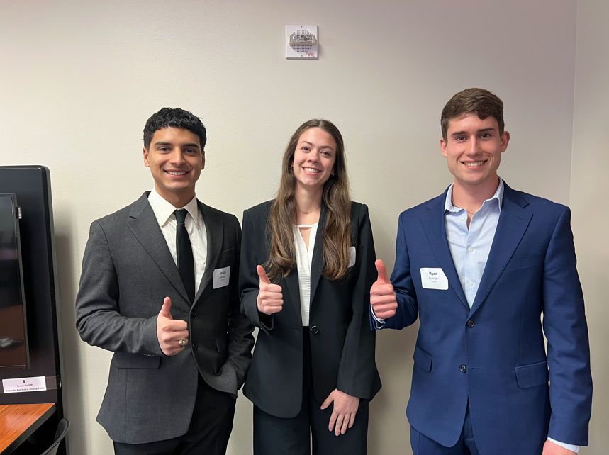 Three individuals stand against a blank white wall. They are each smiling and holding up their thumbs. 