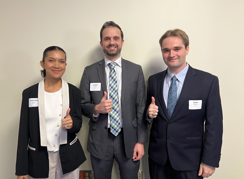 Three people wearing business professional outfits stand against a white wall. They are each smiling and holding up their thumbs. 