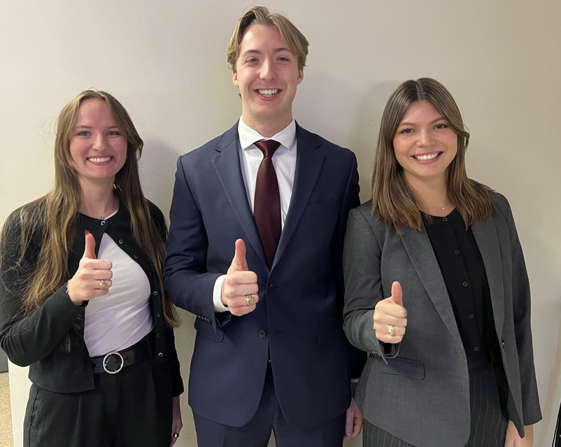 Three students stand against a blank white wall smiling. They are each holding a thumbs-up. 