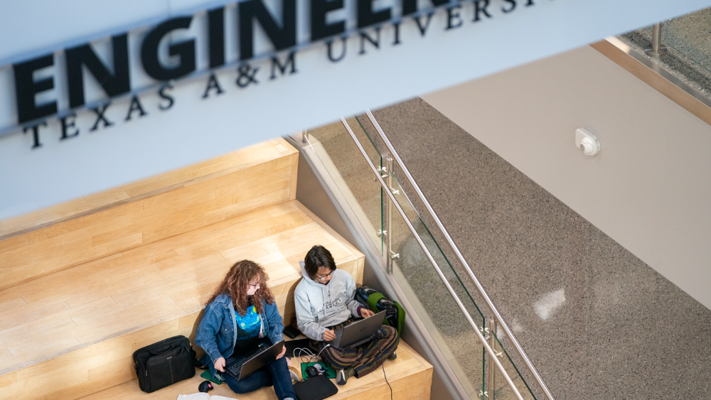Two students sit on a set of steps indoors. They are studying with laptops and notebooks surrounding them. 