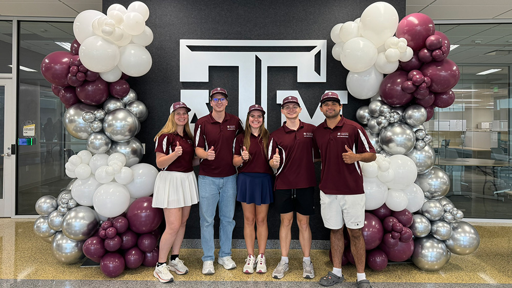 4 ZLP students standing in front of a large Texas A&amp;M logo in the Zachry Engineering Education Complex. There are tall balloon towers, featuring maroon, white, and silver balloons, on either side of the logo, with the students standing in between the two balloon towers.
