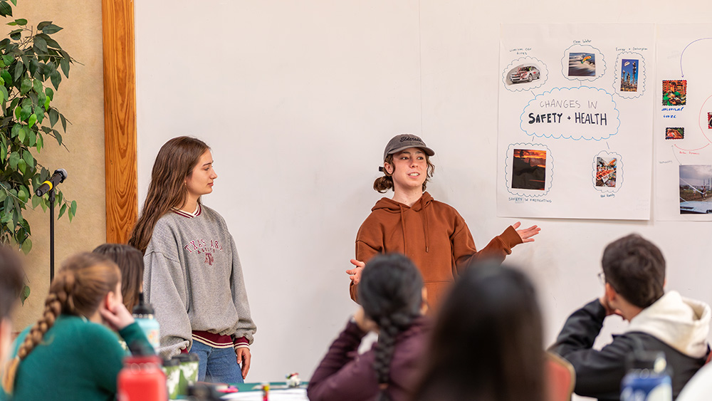 Two students presenting next to a whiteboard while an audience of other students watches.